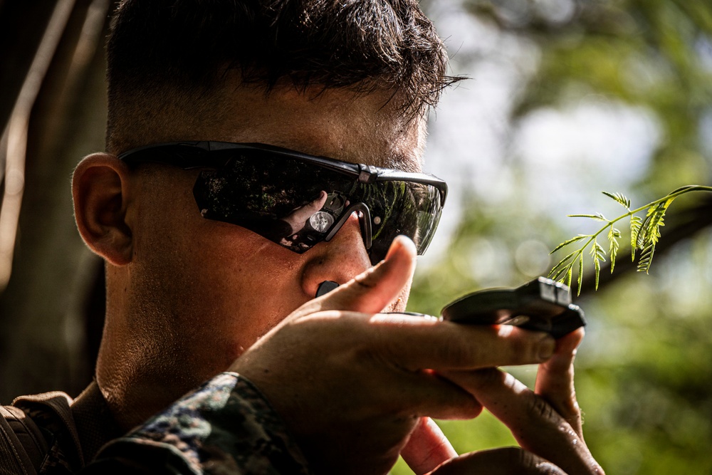 A male individual wearing military camouflage uniform and tactical ballistic sunglasses is aiming a firearm in an outdoor wooded environment. The close-up shot captures him in a shooting stance with a weapon raised to eye level. Green foliage is visible in the foreground and background, suggesting a field or training environment.