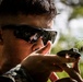 A male individual wearing military camouflage uniform and tactical ballistic sunglasses is aiming a firearm in an outdoor wooded environment. The close-up shot captures him in a shooting stance with a weapon raised to eye level. Green foliage is visible in the foreground and background, suggesting a field or training environment.