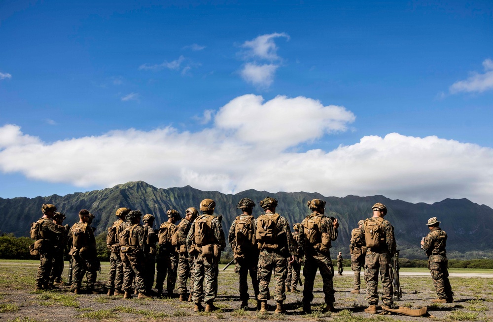 A group of military personnel in full combat gear and camouflage uniforms are assembled on an open field, facing away from the camera toward a figure who appears to be addressing them. The setting features dramatic green mountain ridgelines in the background under a bright blue sky with scattered clouds, consistent with a tropical or Pacific island environment. The scene appears to depict a pre-mission briefing or formation during a training exercise.