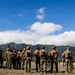 A group of military personnel in full combat gear and camouflage uniforms are assembled on an open field, facing away from the camera toward a figure who appears to be addressing them. The setting features dramatic green mountain ridgelines in the background under a bright blue sky with scattered clouds, consistent with a tropical or Pacific island environment. The scene appears to depict a pre-mission briefing or formation during a training exercise.