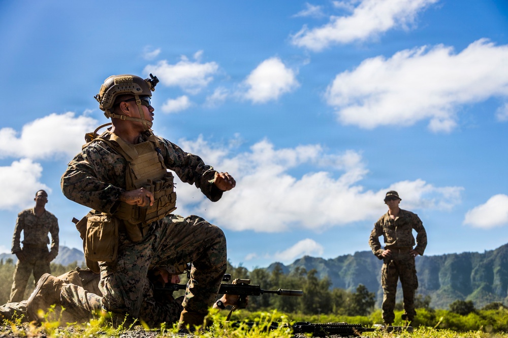 Military personnel in camouflage uniforms are conducting what appears to be a field training exercise in an outdoor range environment. The foreground soldier is kneeling in a tactical position with a rifle on the ground, appearing to give instructions or demonstrate a technique. The setting features lush green mountains and a partly cloudy blue sky in the background, consistent with a tropical or Pacific island location.