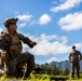 Military personnel in camouflage uniforms are conducting what appears to be a field training exercise in an outdoor range environment. The foreground soldier is kneeling in a tactical position with a rifle on the ground, appearing to give instructions or demonstrate a technique. The setting features lush green mountains and a partly cloudy blue sky in the background, consistent with a tropical or Pacific island location.