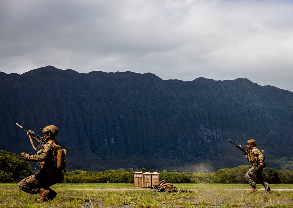Two military personnel in full combat gear are conducting a tactical training exercise on an open field, with one soldier in a crouching position and another running. The scene takes place against a dramatic backdrop of steep, heavily eroded mountain ridgelines consistent with the Ko'olau Range in Hawaii. Portable toilets and a prone figure are visible in the mid-ground, indicating a structured training environment.
