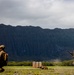 Two military personnel in full combat gear are conducting a tactical training exercise on an open field, with one soldier in a crouching position and another running. The scene takes place against a dramatic backdrop of steep, heavily eroded mountain ridgelines consistent with the Ko'olau Range in Hawaii. Portable toilets and a prone figure are visible in the mid-ground, indicating a structured training environment.