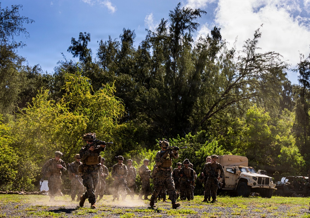 A group of military personnel in camouflage uniforms are conducting what appears to be a field training exercise in a tropical or subtropical outdoor environment. Several soldiers are moving in formation, with at least two carrying shoulder-mounted weapons systems. A tan/beige military tactical vehicle (JLTV or similar) is visible on the right side of the frame.