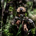 A soldier in full combat gear is positioned in a dense jungle or forest environment, aiming a rifle equipped with an optical scope. The individual is wearing camouflage uniform, a tactical helmet with night-vision device mount, and appears to be in a patrol or reconnaissance posture. The setting suggests a military training exercise or operational deployment in a tropical woodland environment.