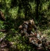 The image shows military personnel in woodland camouflage uniforms operating in a dense jungle or forest environment. One service member appears to be assisting or treating another who is on the ground, while a third moves through the vegetation in the background. The scene appears to depict a combat casualty care drill or field training exercise.