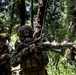 Two military personnel in woodland camouflage uniforms are conducting a patrol or tactical exercise in a dense forested environment. The foreground soldier is aiming a rifle equipped with a suppressor and optical scope, while the second soldier stands in a security posture behind. The setting appears to be a training exercise based on the controlled environment and equipment configuration.