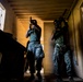 Two armed individuals wearing digital camouflage uniforms are conducting what appears to be a military or law enforcement training exercise inside a building. They are moving through a corridor or room in a tactical formation, with weapons raised in a ready position. The low-angle, high-contrast lighting creates a dramatic visual effect.
