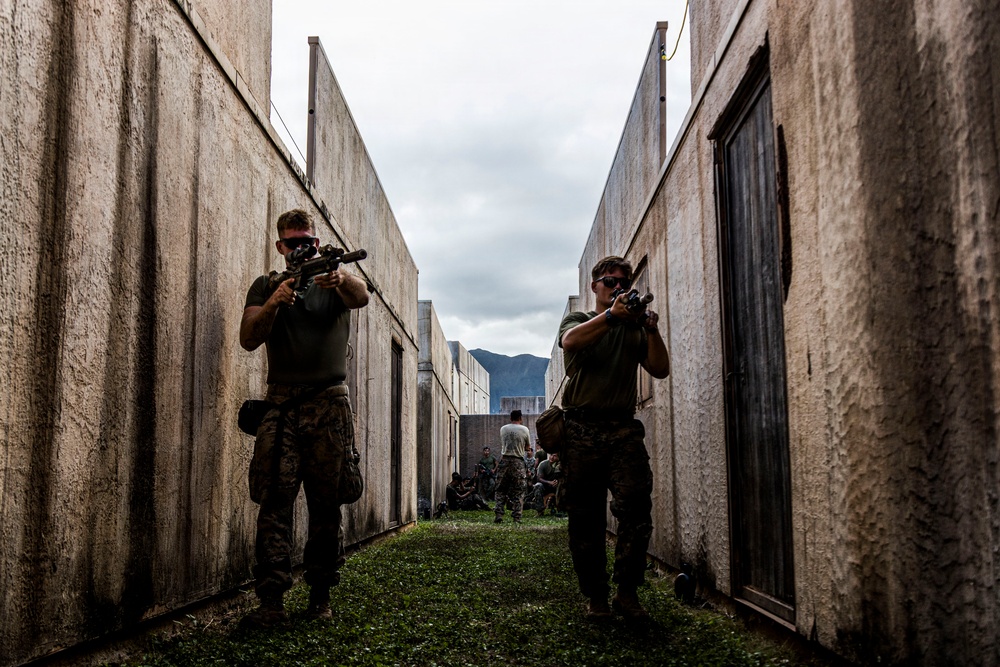 Two armed personnel in military-style clothing are moving tactically through a narrow corridor between concrete walls, with weapons raised in a ready position. The setting appears to be a military training facility or mock urban environment, with additional personnel visible in the background. Mountains and an overcast sky are visible at the end of the corridor.