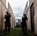 Two armed personnel in military-style clothing are moving tactically through a narrow corridor between concrete walls, with weapons raised in a ready position. The setting appears to be a military training facility or mock urban environment, with additional personnel visible in the background. Mountains and an overcast sky are visible at the end of the corridor.