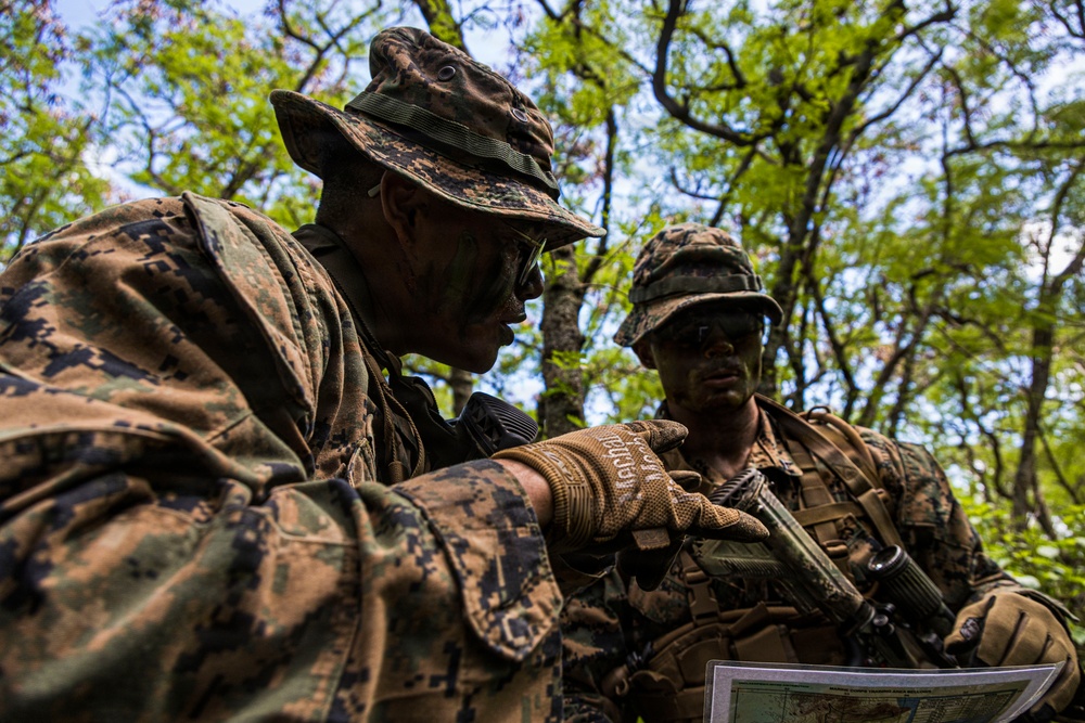 Two military personnel in woodland camouflage uniforms are conducting map reading or navigation planning in a forested environment. Both individuals have camouflage face paint applied and are wearing tactical gear including body armor and boonie hats. One soldier is pointing at a printed map while the other observes, suggesting a patrol briefing or route planning activity.