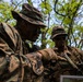 Two military personnel in woodland camouflage uniforms are conducting map reading or navigation planning in a forested environment. Both individuals have camouflage face paint applied and are wearing tactical gear including body armor and boonie hats. One soldier is pointing at a printed map while the other observes, suggesting a patrol briefing or route planning activity.