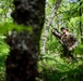 A military service member in woodland camouflage uniform navigates through dense forest terrain. The individual appears to be shouting or calling out while moving through thick undergrowth and trees. The setting suggests a field training exercise or patrol operation in a lush, green forested environment.