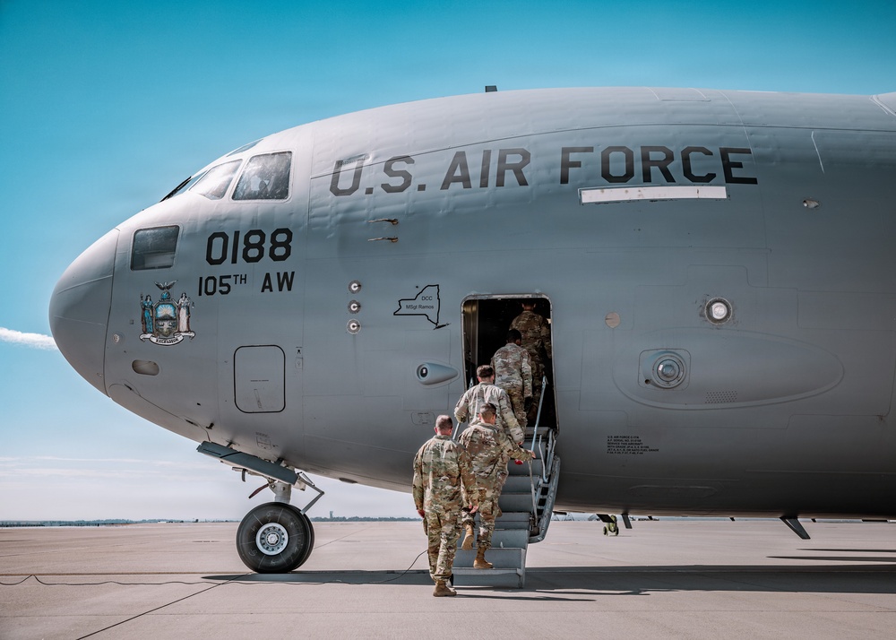 Military personnel in camouflage uniforms are boarding a U.S. Air Force aircraft on a tarmac under clear blue skies. The aircraft is marked with tail number 0188 and assigned to the 105th Airlift Wing, featuring the New York State seal. Four service members are visible ascending a mobile staircase into the forward entry door of the aircraft.