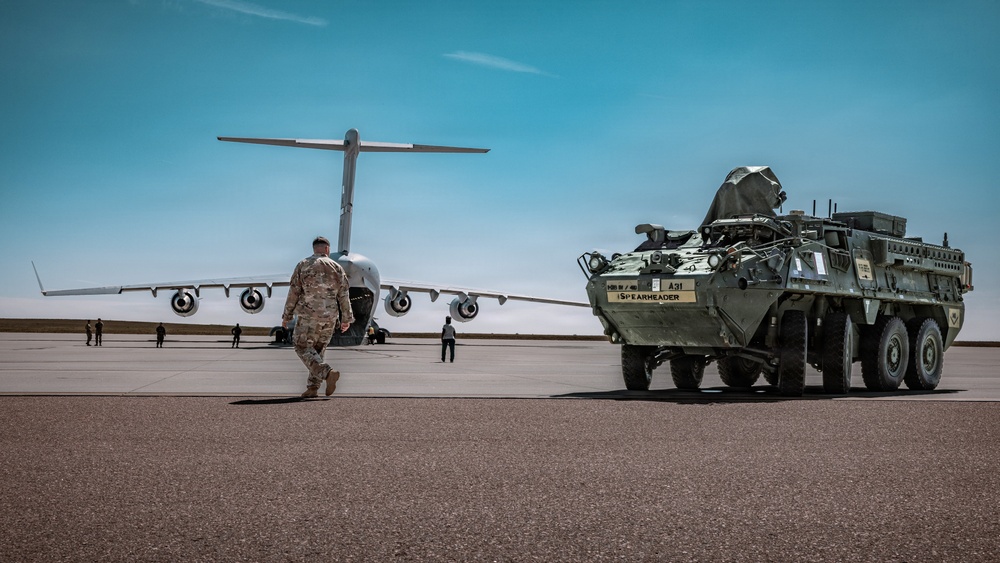 A military airfield scene showing a large transport aircraft (consistent with a C-17 Globemaster III) on a tarmac alongside an armored wheeled vehicle marked 'A31 SPEARHEADER'. A soldier in camouflage uniform walks toward the aircraft, with several other personnel visible in the background. The scene appears to depict a military deployment or equipment loading/unloading operation.