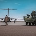 A military airfield scene showing a large transport aircraft (consistent with a C-17 Globemaster III) on a tarmac alongside an armored wheeled vehicle marked 'A31 SPEARHEADER'. A soldier in camouflage uniform walks toward the aircraft, with several other personnel visible in the background. The scene appears to depict a military deployment or equipment loading/unloading operation.