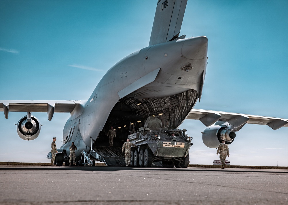 A large military transport aircraft, identifiable as a C-5 Galaxy (tail number 10188), has its nose visor raised to allow loading or unloading of a wheeled armored vehicle. The armored vehicle, marked 'A31' and labeled '1SPEARHEADER', appears to be a Stryker infantry carrier vehicle being driven onto or off the aircraft's cargo ramp. Several military personnel in camouflage uniforms are supervising and guiding the operation on the tarmac.