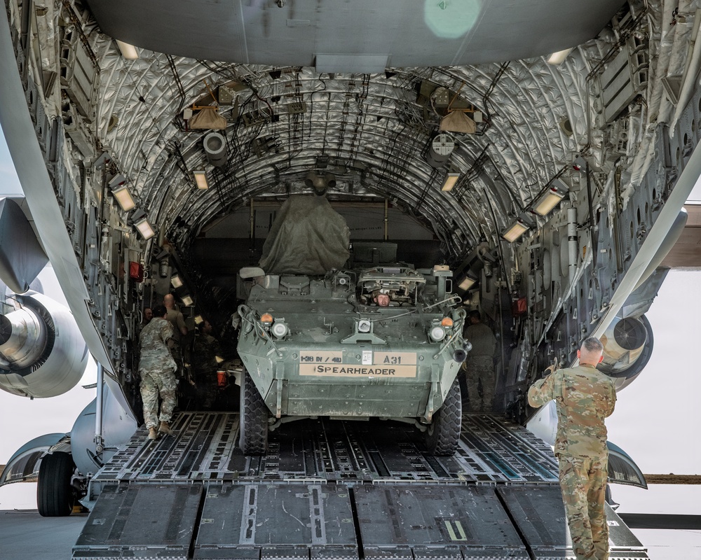 A U.S. Army Stryker armored fighting vehicle is being loaded into the cargo bay of a large military transport aircraft, likely a C-17 Globemaster III. The vehicle bears the markings 'A31', '1-28 IN / 4ID', and 'SPEARHEADER', identifying it as belonging to the 1st Battalion, 28th Infantry Regiment, 4th Infantry Division. Several soldiers in camouflage uniforms are guiding and supervising the loading operation on the aircraft's rear ramp.
