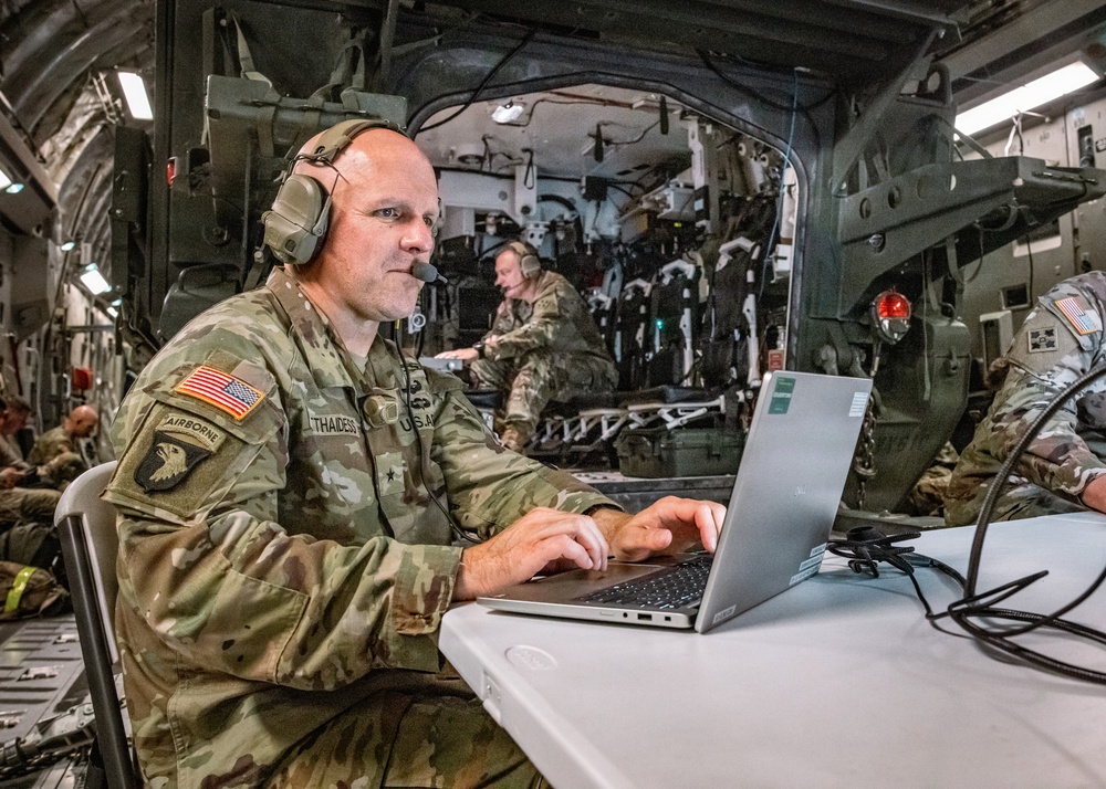 A U.S. Army soldier wearing a headset is working on a laptop computer at a folding table inside what appears to be a military transport aircraft. The soldier wears a 101st Airborne Division patch and a name tape reading 'THAIDES' with a general officer star visible. Several other uniformed personnel are visible in the background of the aircraft's cargo bay.