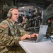 A U.S. Army soldier wearing a headset is working on a laptop computer at a folding table inside what appears to be a military transport aircraft. The soldier wears a 101st Airborne Division patch and a name tape reading 'THAIDES' with a general officer star visible. Several other uniformed personnel are visible in the background of the aircraft's cargo bay.