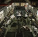 The interior of a large military transport aircraft, likely a C-17 Globemaster III, is shown loaded with military personnel and at least one armored vehicle covered with a tarpaulin. Soldiers are seated along the sidewall jump seats on both port and starboard sides of the fuselage. The cargo hold is fully utilized with equipment, gear bags, and what appears to be a wheeled armored vehicle secured to the cargo floor.