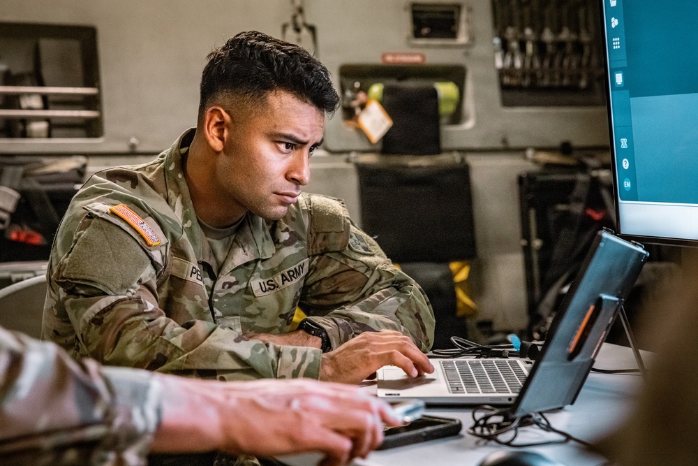 A U.S. Army soldier in OCP (Operational Camouflage Pattern) uniform is focused on working at a laptop computer in what appears to be a military workspace or vehicle interior. The soldier's name tape reads 'PE[R/N]...' and the 'U.S. ARMY' tape is clearly visible along with an American flag patch on the left shoulder. A second person's arm is partially visible in the foreground, and a large monitor is visible to the right.