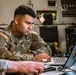 A U.S. Army soldier in OCP (Operational Camouflage Pattern) uniform is focused on working at a laptop computer in what appears to be a military workspace or vehicle interior. The soldier's name tape reads 'PE[R/N]...' and the 'U.S. ARMY' tape is clearly visible along with an American flag patch on the left shoulder. A second person's arm is partially visible in the foreground, and a large monitor is visible to the right.