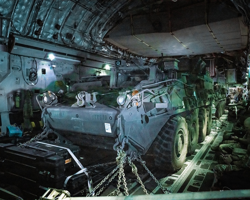 The interior of a large military transport aircraft, likely a C-17 Globemaster III, loaded with at least two Stryker armored fighting vehicles secured with chains and tie-down straps. The vehicles are chained to the aircraft floor with heavy-duty restraints, and military personnel are seated along the right side of the fuselage. An American flag is visible on the right side of the cargo hold.