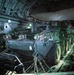 The interior of a large military transport aircraft, likely a C-17 Globemaster III, loaded with at least two Stryker armored fighting vehicles secured with chains and tie-down straps. The vehicles are chained to the aircraft floor with heavy-duty restraints, and military personnel are seated along the right side of the fuselage. An American flag is visible on the right side of the cargo hold.