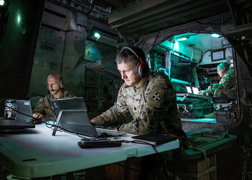 Three military personnel in camouflage uniforms are working inside what appears to be a tactical command vehicle or mobile operations center. The central figure, identified by a name tape as 'KRIEGER,' is focused on a laptop while wearing red and black headphones, with a 4th Infantry Division patch visible on their shoulder. The scene is illuminated by artificial lighting in a dimly lit, equipment-filled military vehicle interior.