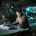 Three military personnel in camouflage uniforms are working inside what appears to be a tactical command vehicle or mobile operations center. The central figure, identified by a name tape as 'KRIEGER,' is focused on a laptop while wearing red and black headphones, with a 4th Infantry Division patch visible on their shoulder. The scene is illuminated by artificial lighting in a dimly lit, equipment-filled military vehicle interior.