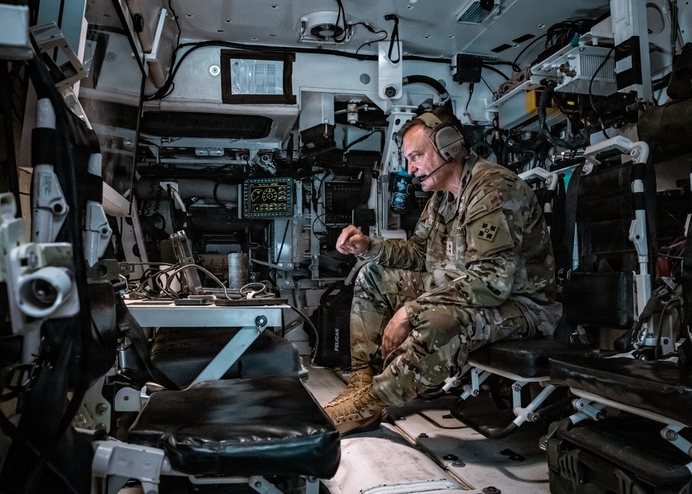 A U.S. Army soldier in MultiCam/OCP camouflage uniform is seated inside what appears to be a military command vehicle or armored personnel carrier. He is wearing a communications headset and appears to be speaking, possibly conducting or participating in a briefing or radio communication. The interior is densely packed with electronic equipment, displays, cables, and military hardware.