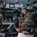 A U.S. Army soldier in MultiCam/OCP camouflage uniform is seated inside what appears to be a military command vehicle or armored personnel carrier. He is wearing a communications headset and appears to be speaking, possibly conducting or participating in a briefing or radio communication. The interior is densely packed with electronic equipment, displays, cables, and military hardware.