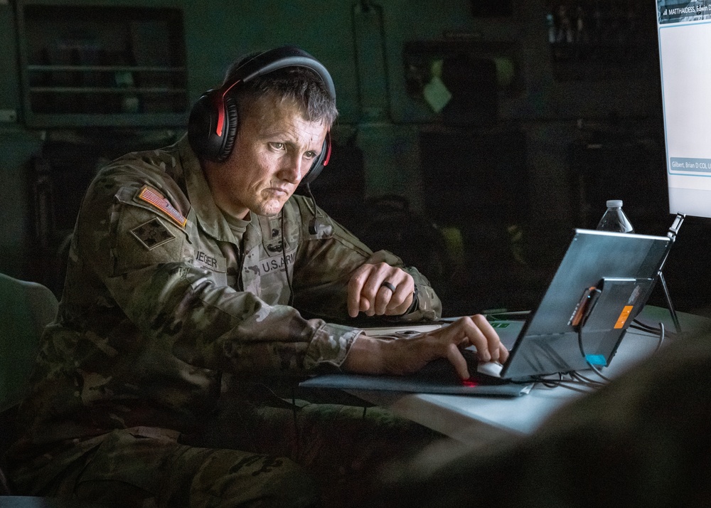 A U.S. Army soldier wearing camouflage uniform and red-accented black headphones is working intently at a laptop computer in a dimly lit environment. The soldier's name tape reads 'IEGER' and the uniform displays a U.S. flag patch on the left shoulder. A secondary monitor is visible in the background displaying what appears to be a personnel or communications interface.