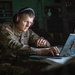 A U.S. Army soldier wearing camouflage uniform and red-accented black headphones is working intently at a laptop computer in a dimly lit environment. The soldier's name tape reads 'IEGER' and the uniform displays a U.S. flag patch on the left shoulder. A secondary monitor is visible in the background displaying what appears to be a personnel or communications interface.