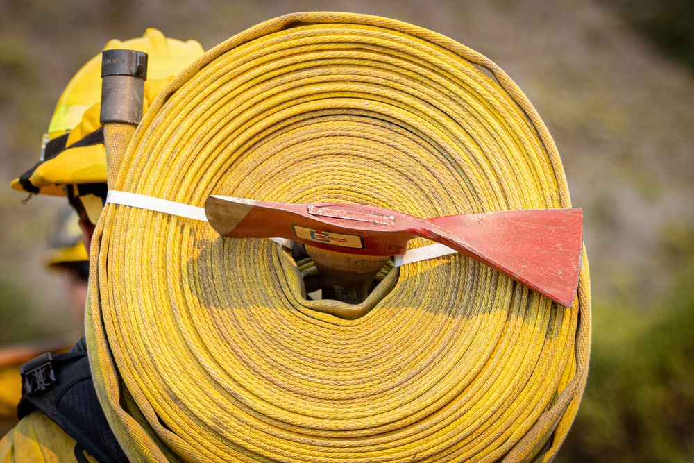 A firefighter is carrying a large coiled yellow fire hose on their back or shoulder. A red-painted hand tool, likely a fire swatter or flap tool used in wildland firefighting, is secured across the hose roll with a white strap. The firefighter is wearing standard wildland firefighting gear including a yellow Nomex jacket and helmet.