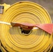 A firefighter is carrying a large coiled yellow fire hose on their back or shoulder. A red-painted hand tool, likely a fire swatter or flap tool used in wildland firefighting, is secured across the hose roll with a white strap. The firefighter is wearing standard wildland firefighting gear including a yellow Nomex jacket and helmet.