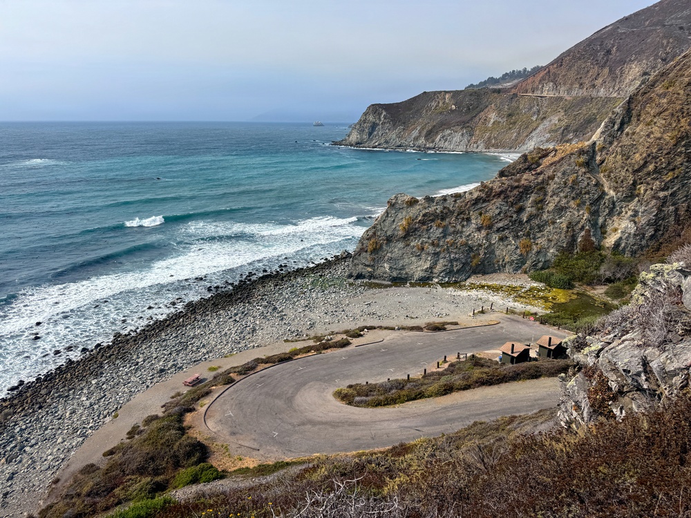 A dramatic coastal scene featuring a small, empty paved parking area or turnout situated at the base of steep rocky cliffs along a rugged shoreline. The turquoise-blue ocean with breaking waves dominates the left side of the image, while layered sedimentary cliffs extend into the background. A winding road is faintly visible carved into the upper cliffs, suggesting this is likely along California's Highway 1 near Big Sur.