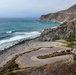 A dramatic coastal scene featuring a small, empty paved parking area or turnout situated at the base of steep rocky cliffs along a rugged shoreline. The turquoise-blue ocean with breaking waves dominates the left side of the image, while layered sedimentary cliffs extend into the background. A winding road is faintly visible carved into the upper cliffs, suggesting this is likely along California's Highway 1 near Big Sur.