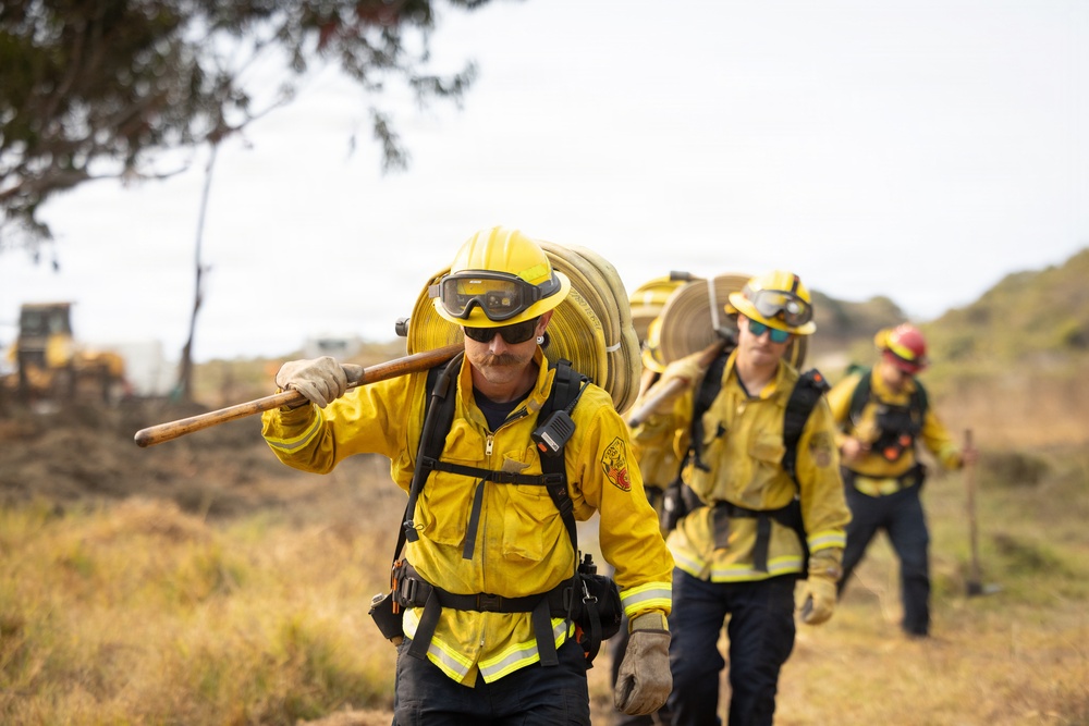 A team of wildland firefighters in yellow protective gear are walking across dry, grassy terrain while carrying fire hoses and hand tools. The crew appears to be moving toward or away from a fire line, with heavy equipment visible in the background. The image captures the physical demands of wildfire suppression operations.