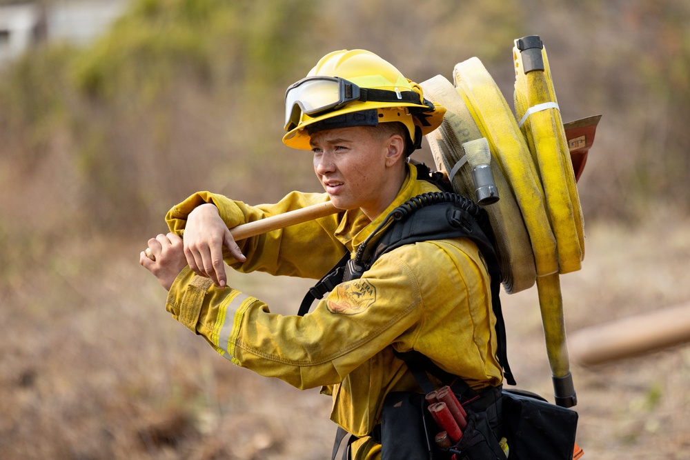 A young firefighter in full wildland firefighting gear is photographed in an outdoor, dry brush environment. They are carrying coiled yellow fire hoses over their shoulder along with a hand tool, suggesting active deployment or post-suppression work. The image is a close-up portrait-style shot that emphasizes the subject's equipment and expression.