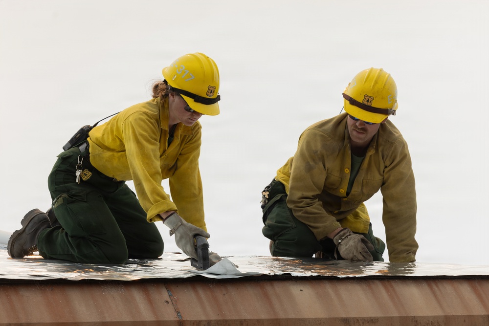 Two U.S. Forest Service workers are kneeling on what appears to be a metal roof or surface, working with tools on a reflective material. Both are wearing standard wildland firefighter gear including yellow Nomex shirts, green pants, yellow hard hats with USFS shields, safety glasses, and work gloves. They appear to be applying or securing a fire-resistant reflective wrap or barrier material.