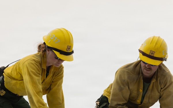 Two U.S. Forest Service workers are kneeling on what appears to be a metal roof or surface, working with tools on a reflective material. Both are wearing standard wildland firefighter gear including yellow Nomex shirts, green pants, yellow hard hats with USFS shields, safety glasses, and work gloves. They appear to be applying or securing a fire-resistant reflective wrap or barrier material.