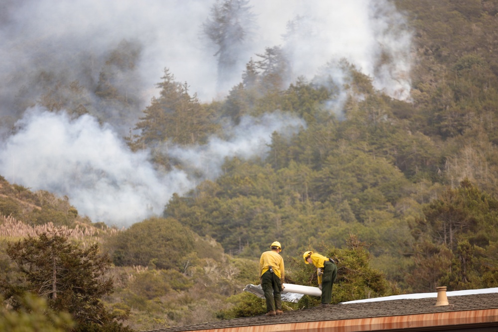 Two wildland firefighters in yellow shirts and green pants are working on a residential rooftop, appearing to deploy protective fire-resistant wrap or sheeting to defend the structure. A large wildfire is actively burning on a forested hillside directly behind them, with heavy white and gray smoke billowing through the tree canopy. The scene depicts a structure protection operation with the fire posing an imminent threat to the building.