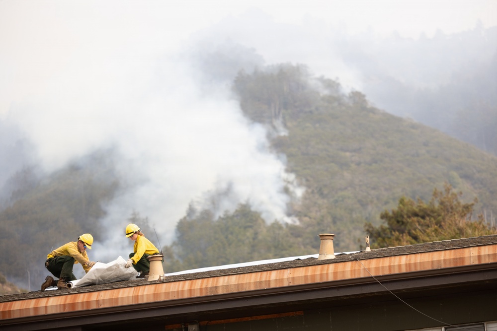 Two firefighters in yellow protective gear are on the roof of a structure, deploying what appears to be fire-resistant wrap or protective sheeting to defend the building from an approaching wildfire. A heavily forested hillside or mountain is visible in the background, with multiple columns of smoke rising from active fire burning through the vegetation. The scene depicts active structure protection operations during a wildfire event.