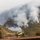 Two firefighters in yellow protective gear are positioned on a residential rooftop, carrying a fire hose to defend the structure from an approaching wildfire. A heavily forested hillside behind them is actively burning, with large plumes of gray-white smoke rising from multiple ignition points. The scene depicts a defensive firefighting operation to protect a home from an encroaching wildland fire.
