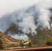 Two firefighters in yellow protective gear are positioned on a residential rooftop, carrying a fire hose to defend the structure from an approaching wildfire. A heavily forested hillside behind them is actively burning, with large plumes of gray-white smoke rising from multiple ignition points. The scene depicts a defensive firefighting operation to protect a home from an encroaching wildland fire.