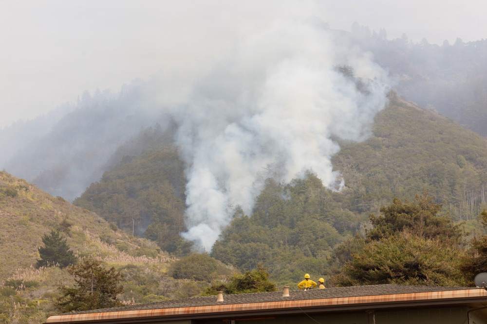 A wildfire burns on a heavily vegetated hillside, producing large plumes of white-gray smoke that obscure the upper portion of the mountain. In the foreground, three firefighters wearing yellow protective gear and hard hats are visible standing on the roof of a building, apparently monitoring the fire situation. The scene suggests an active wildfire response with structures potentially at risk.