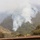 A wildfire burns on a heavily vegetated hillside, producing large plumes of white-gray smoke that obscure the upper portion of the mountain. In the foreground, three firefighters wearing yellow protective gear and hard hats are visible standing on the roof of a building, apparently monitoring the fire situation. The scene suggests an active wildfire response with structures potentially at risk.
